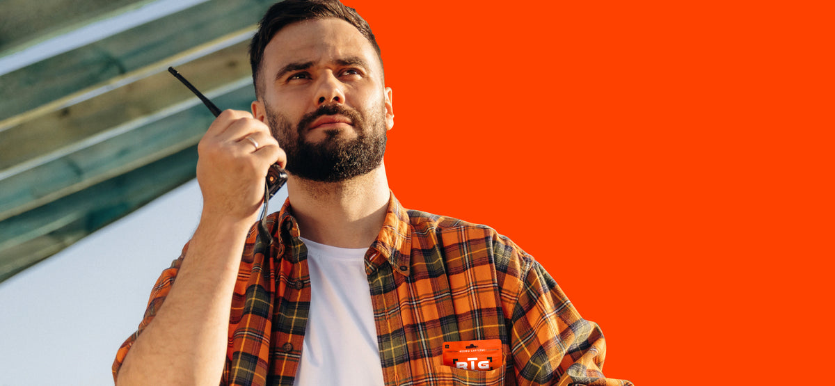 A man with a beard and mustache wearing a brown and orange plaid flannel shirt with a pouch of RIG chews in the pocket over a white t-shirt, holding a walkie-talkie to his ear. He is positioned against a solid bright orange background with a wooden structure visible on the left.